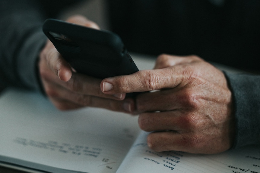 A close up of a man's hands in a dimly lit room, as he makes a call on a mobile phone.