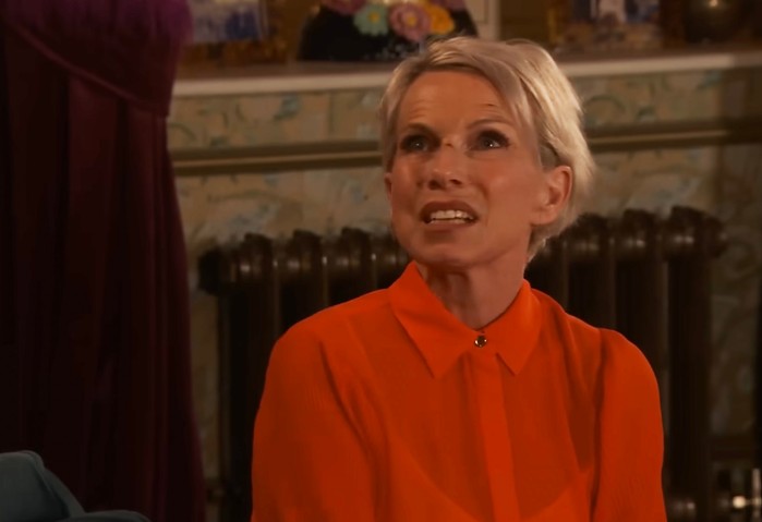 Debbie Webster Debbie Webster looks up towards someone from a sofa. She's wearing an orange shirt and looks exasperated with cuts on her face.