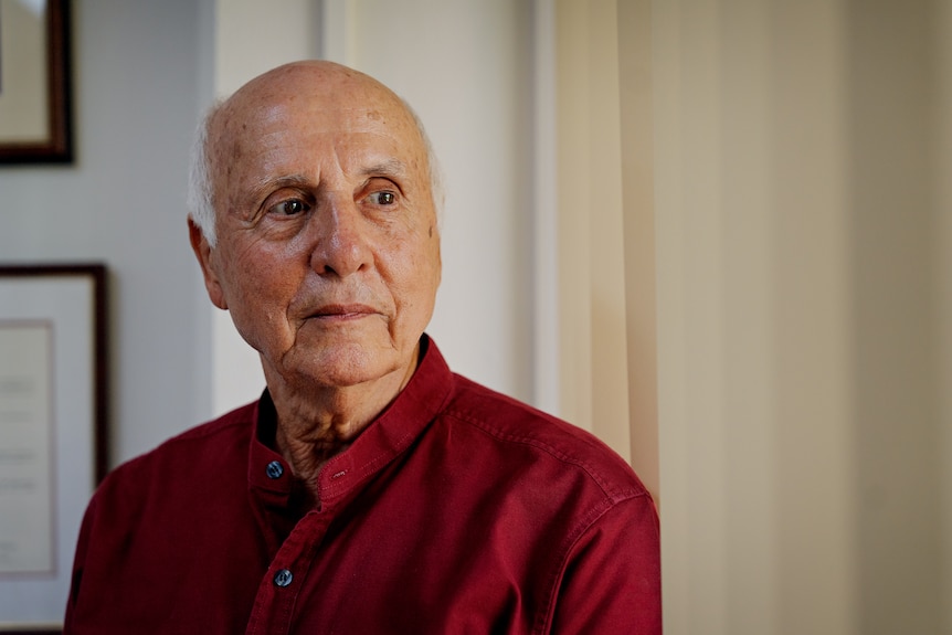 A senior, white man, bald top head, with white side hair looking right of camera, wearing a deep read button-up shirt