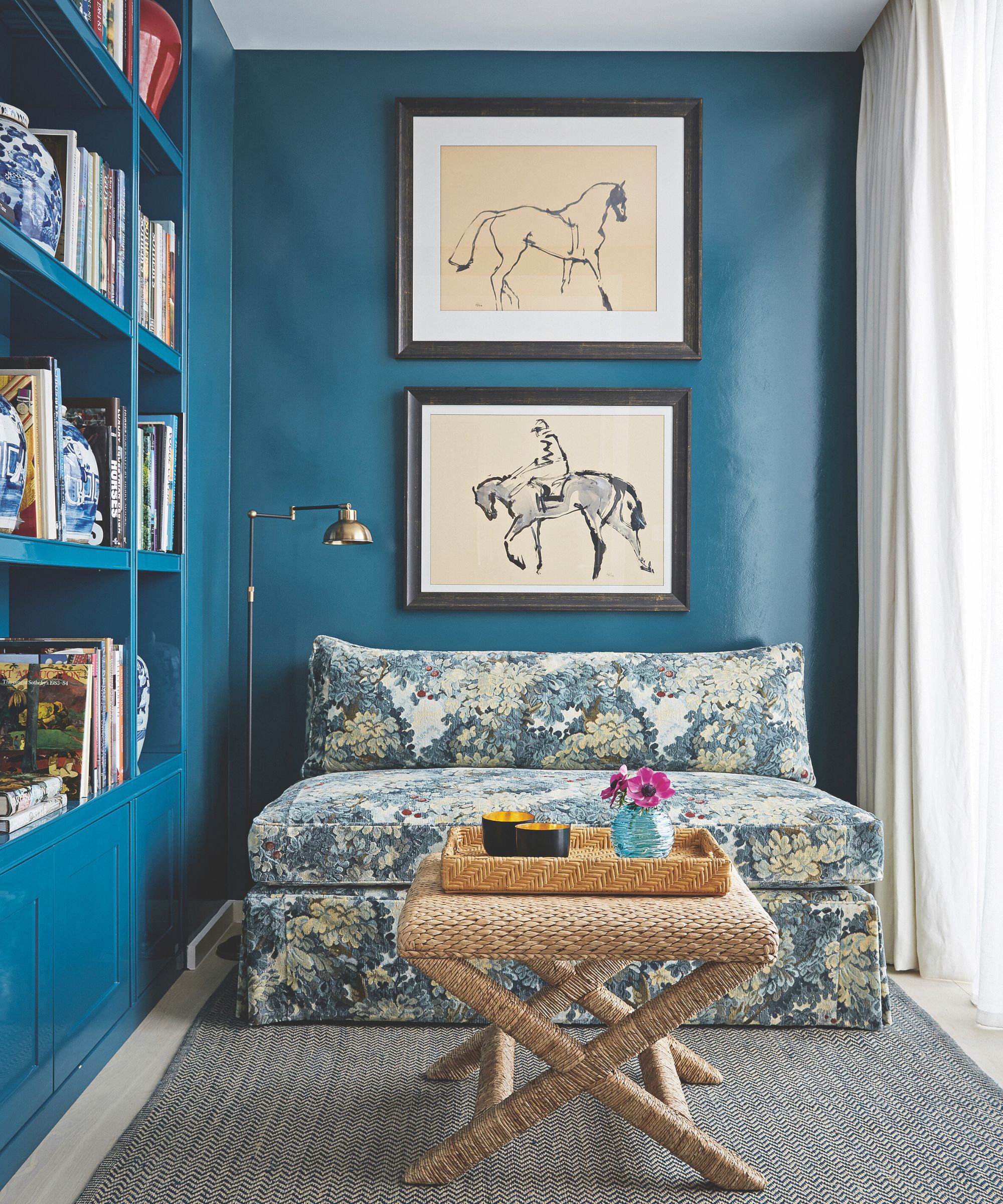 a blue-drenched room with a floral sofa, a woven ottoman, and built-in bookshelves