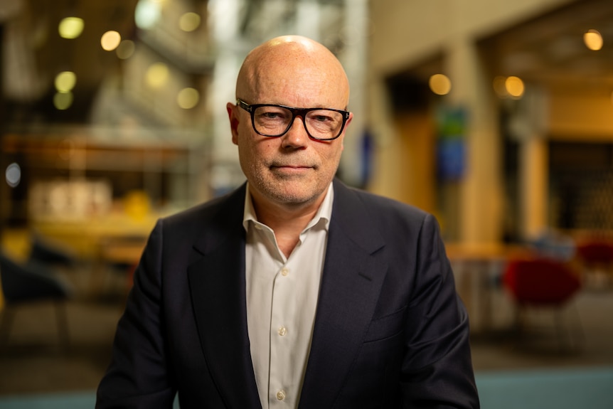 A man in dark rimmed glasses sits at a desk pensively.