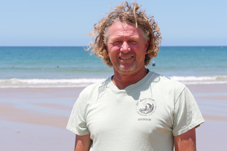 Paul White with blond curly hair, wearing a t-shirt smiling directly at the camera with the beach in the background.