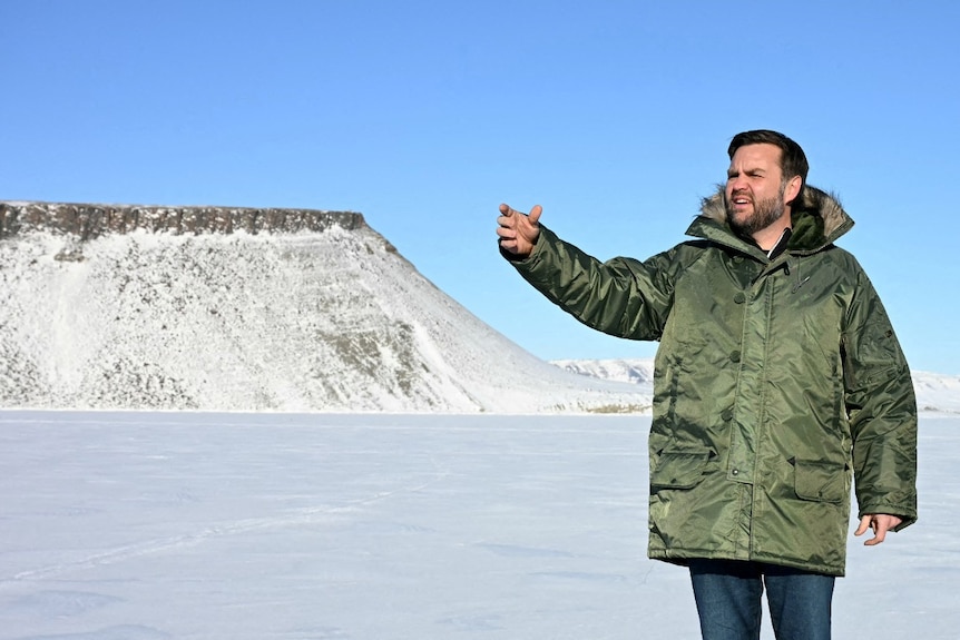 A man in a large coat stands among a snowy landscape with his hand raised.