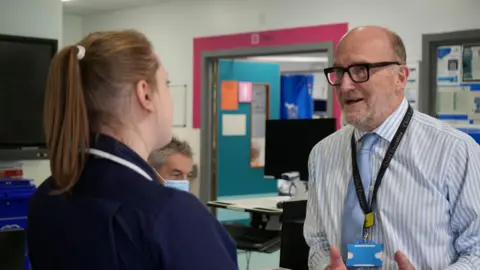 JAMIE NIBLOCK/BBC Nick Hulme stands at the reception desk in the elderly ward at Colchester Hospital talking to the ward matron. She is wearing a navy blue dress with white trim and has her hair pulled back in a pony tail. He is wearing a lanyard around his neck. He has a pale blue and white pinstripe shirt and a blue tie. He is wearing thick plastic black glasses and has a short beard. 