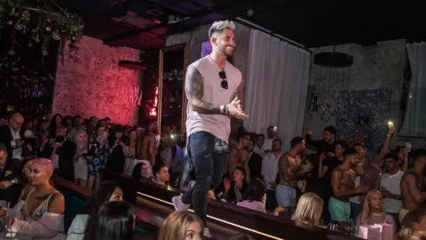 The Couture Club Ross Worswick is walking along a catwalk modelling some of the company's clothing in a buys venue. He is wearing jeans and a pale pink sleeveless top with sunglasses hanging from the neckline. 