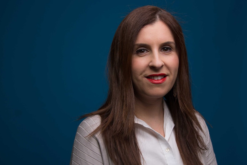 A woman with long brown hair, facing the camera, against a matt blue background.