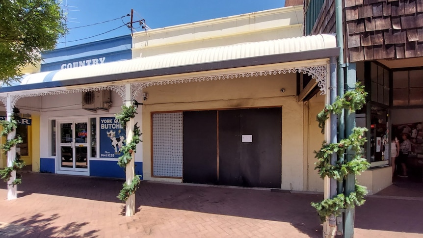 A boarded up terrace shop. Part of the facade is black from smoke or burn marks. Businesses either side are open.