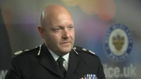 BBC A bald man wearing a black uniform looks to the right of the frame. He is in front of a blurred background with the badge of West Midlands Police in frame.