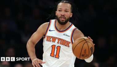 Giannis Antetokounmpo is wearing a green Milwaukee Bucks vest and holding a basketball