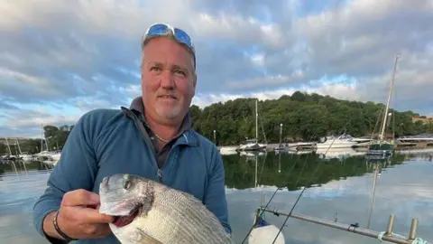 Chris Puncher Chris Puncher stood to the left wearing a blue jumper. He's looking at the camera and holding a large fish. In the background is water and land in the distance. 