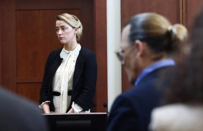 Amber Heard standing in a courtroom wearing a white blouse and dark blazer, appearing attentive. Johnny Depp, blurred in the foreground, with tied-back hair