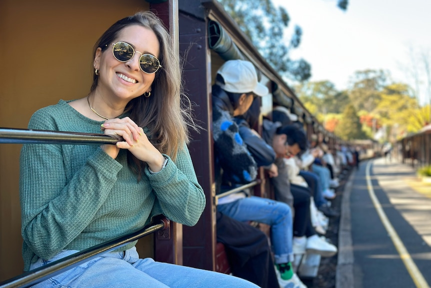 Woman smiles wearing sunglasses, sitting alongside other passengers with legs dangling out of train