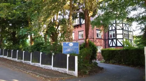 Google A tree and hedge lined drive way leading to a brick building with Victorian style white fascia lined with black wooden beams. A blue sign marks the entrance with 'Norway Lodge' in white letters. 