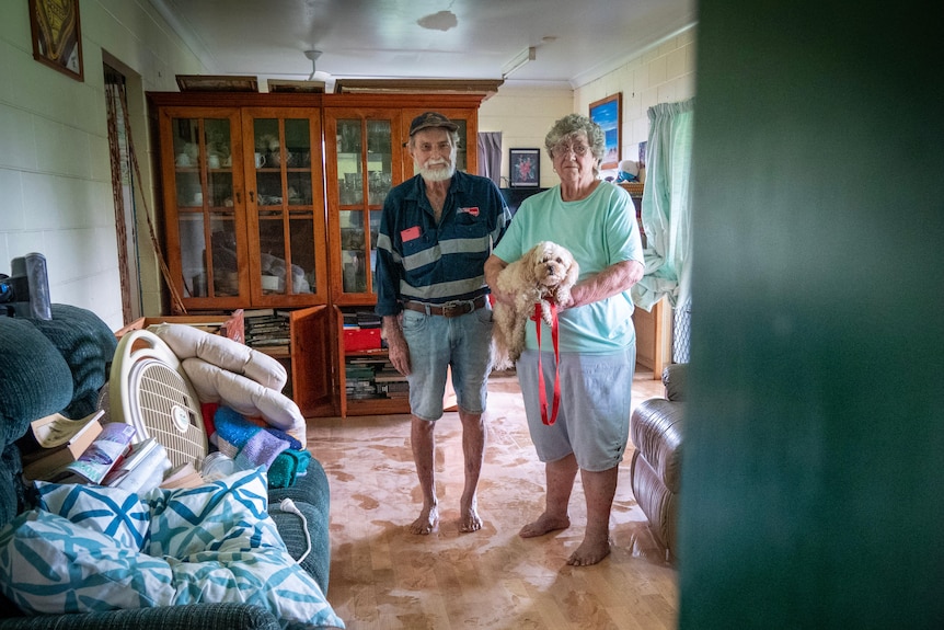 A man and woman stand together in their lounge room with water and mud on the floor.