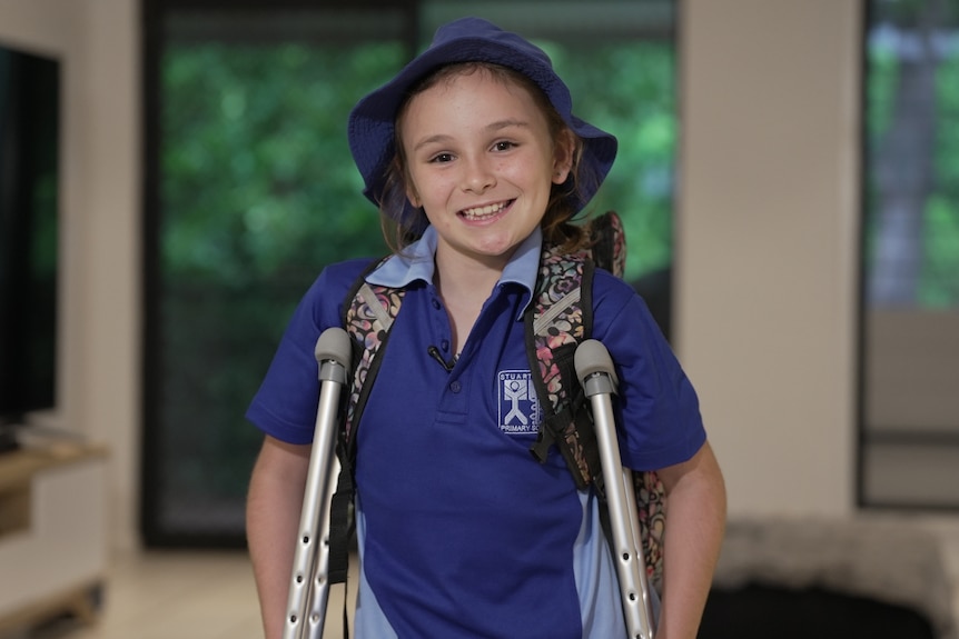 A young girl, dressed in her blue primary school uniform and holding crutches as she smiles at the camera.