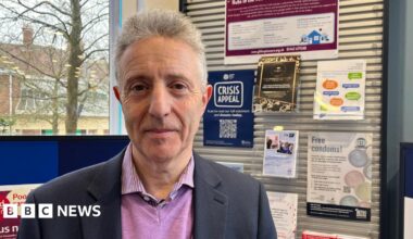 A man with grey hair stands in front of a health notice board in a pharmacy. The posters on the board promote local charities and health advice.