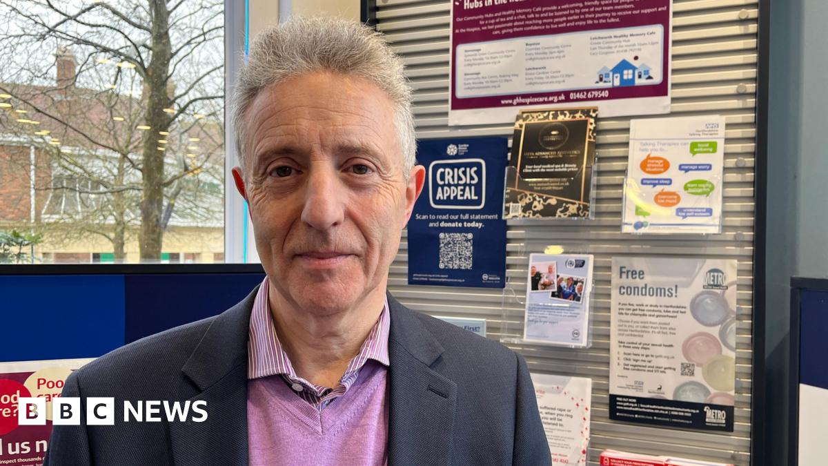 A man with grey hair stands in front of a health notice board in a pharmacy. The posters on the board promote local charities and health advice.