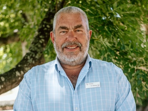 A man with grey hair and beard, wearing an open-necked shirt, standing in front of a tree.