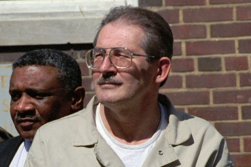 A man with glasses and moustache leaves court with files under his arm, and in handcuffs.