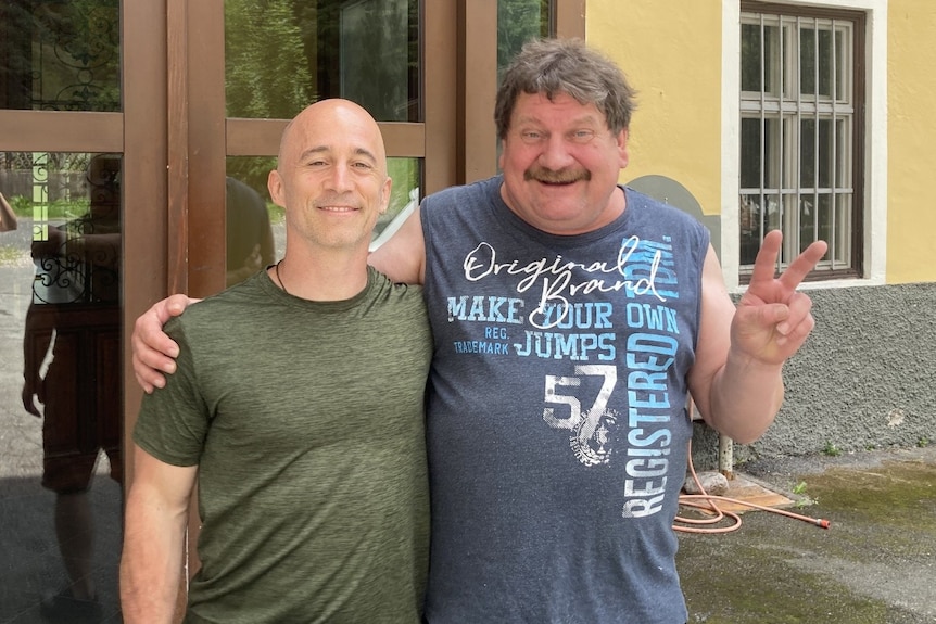 Two men in T-shirts smile while standing arm in arm in front of a house.