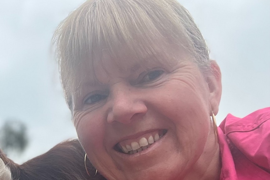 A close up shot of a woman with blonde hair, big hoop earrings and a pink shirt smiling.