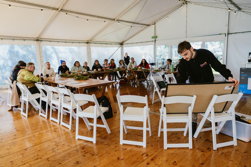 A group of people sit in a semi circle at tables while a young man with short brown hair sets up a board for them to look at.