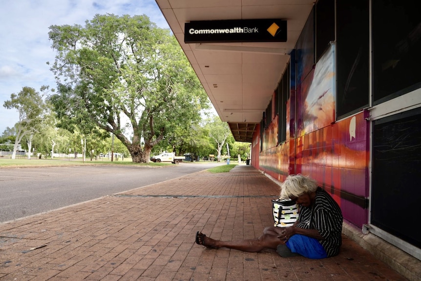old Aboriginal woman sitting on the ground next to Commonwealth bank. 