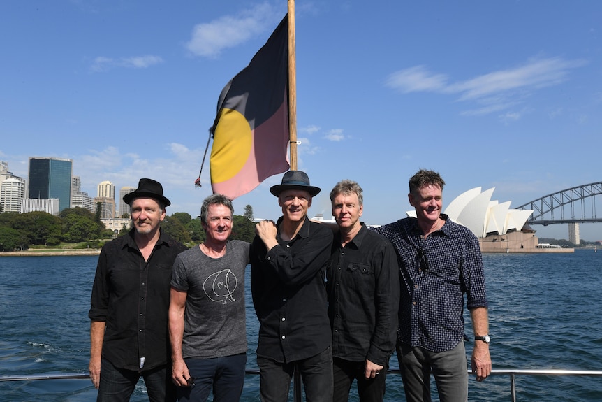 Five men stand in front of Sydney Harbour, an Aboriginal flag flying behind them and the Sydney Opera House in the background.