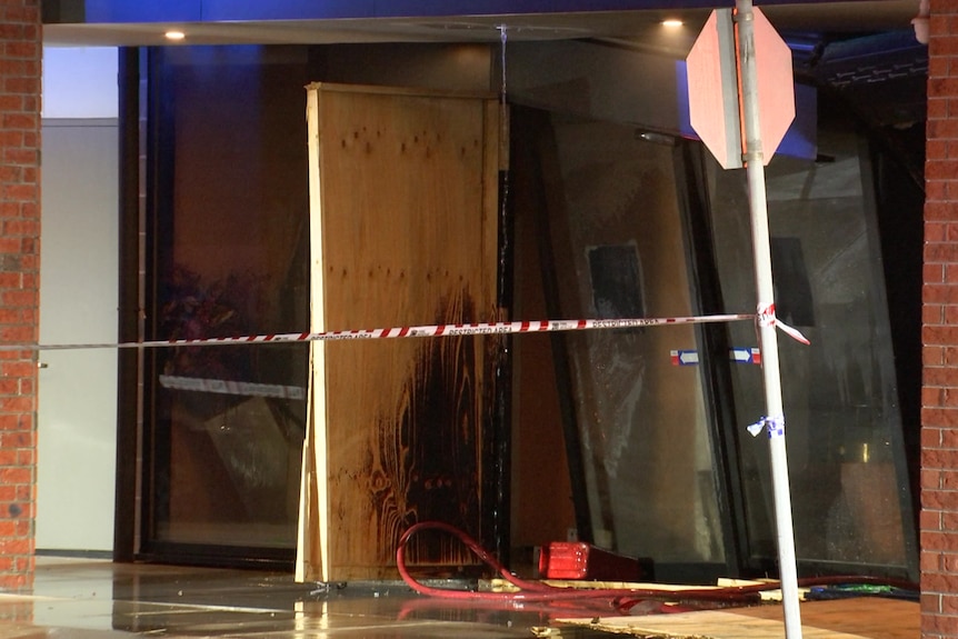 A red jerry can lies on the ground next to a wooden door with a black scorch mark beside damaged glass panels.