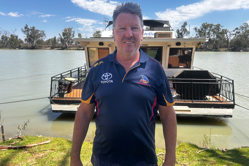 A man in a blue shirt stands in front of a houseboat moored in a muddy river.