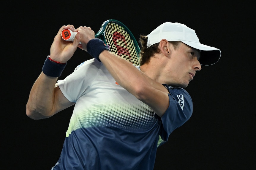 Alex de Minaur swings his racquet behind him. He wears a white cap and a green, blue and white shirt.