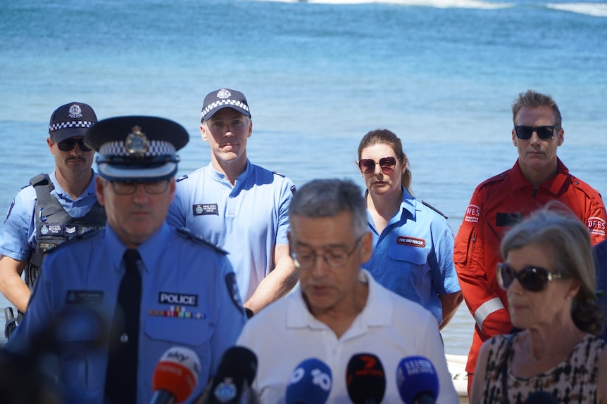 A group of people stand by along a river as two people speak into a mic