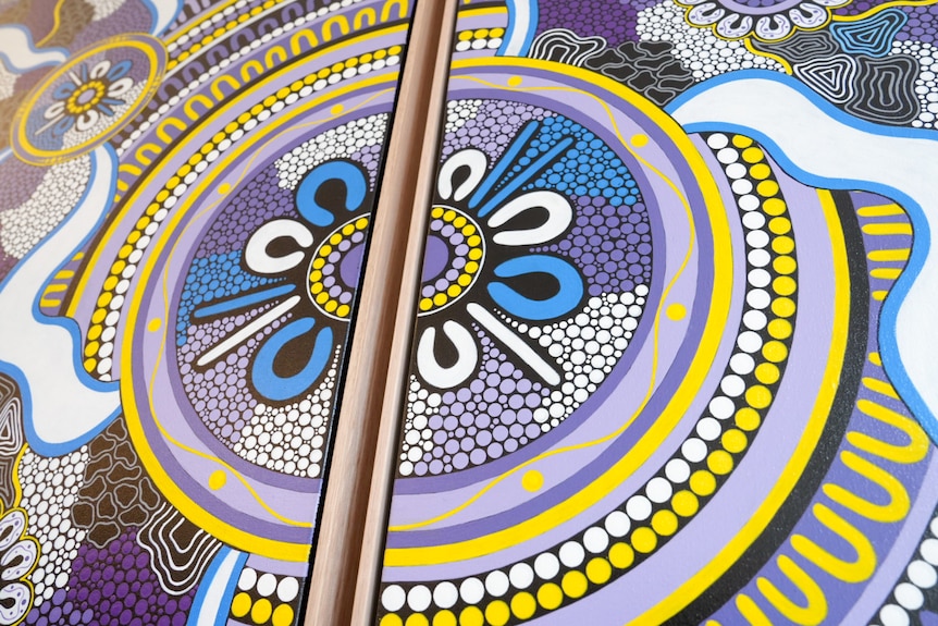 Purple, white and yellow coloured Aboriginal artwork featuring people seated made up of colourful shapes and dots.