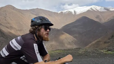 Olly Hargreaves Olly sits on his bike and looks away from the camera. He wears a black bike helmet and sunglasses. He has collar-length blonde hair and a ginger beard. Mountains are visible in the distance.