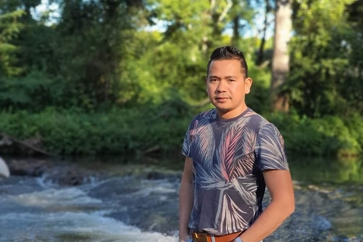 Bunchhay is a young SE Asian man in an grey and pink T-shirt stands in a leafy outdoor setting.