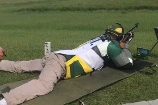 A man in a green, white and yellow shirt lies on the ground aiming a rifle.