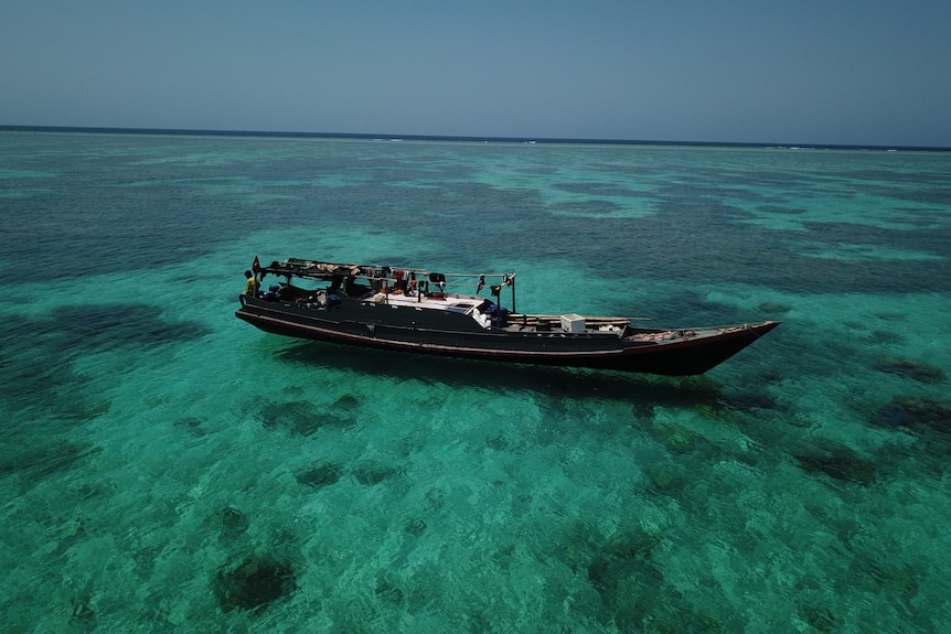 A man sitting on the edge of a long wooden fishing boat, floating on the water on top of a reef.