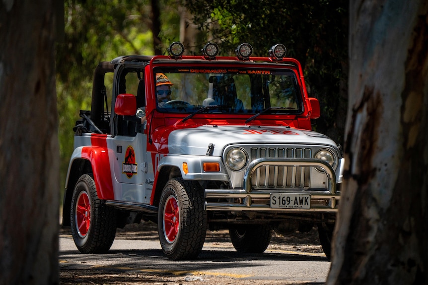 A red and silver jeep similar to the one in the movie Jurassic Park being driven past large trees