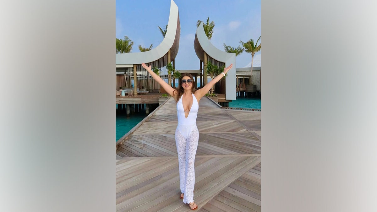 Elizabeth Hurley poses with her arms up on a jetty in the Maldives in a white bathing suit.