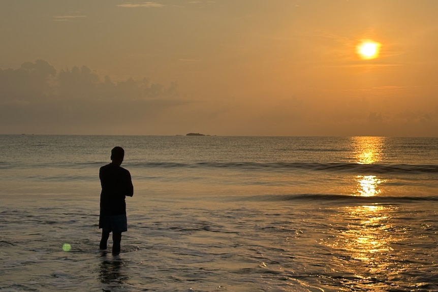 A person standing in water at a beach at sunset.
