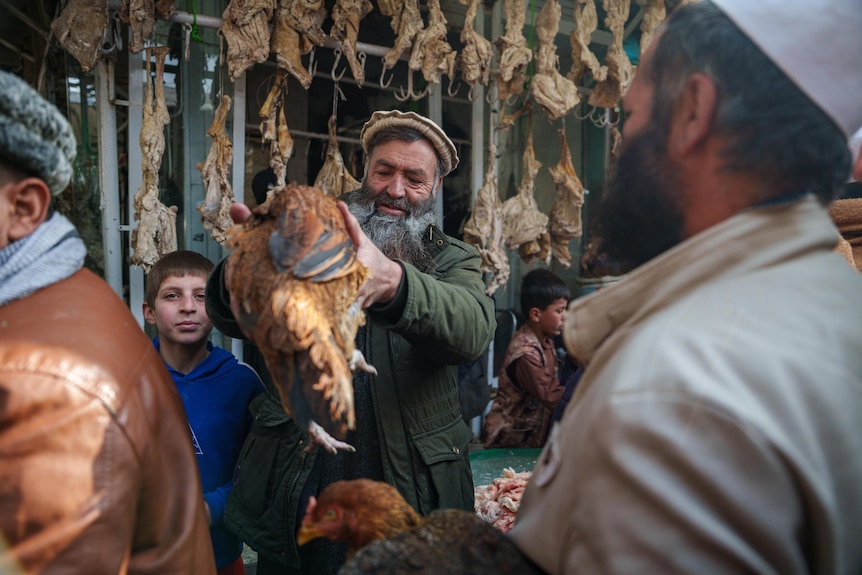A man holds a chicken up. The chicken is alive but drying meats hang in the store behind him.