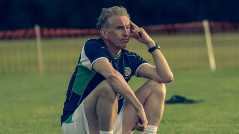 Aidan Monaghan Steve Coogan sitting on the sidelines in a Republic of Ireland training kit looking disconsolate. He has grey hair and has his left elbow leaning on his knee.