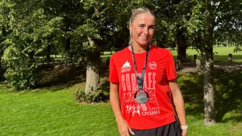 Leah Walsh Leah smiling looking at the camera. She is stood in front of trees and greenery and wears a red Cardiff half marathon finishers t-shirt, a black medal and black running shorts. Leah has blonde hair in a high ponytail. 
