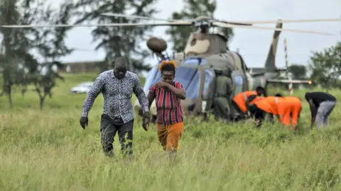 EPA/Shutterstock A man and a women walk away from a military helicopter as members of a rescue teams in orange coats help someone in a stretcher in Mozambique
