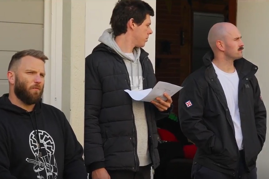 three men stand near a panneled house dressed sloppily.