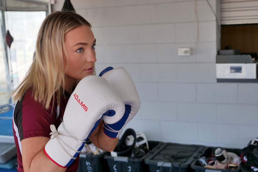 A young woman wearing boxing gloves, looking intensely off-camera