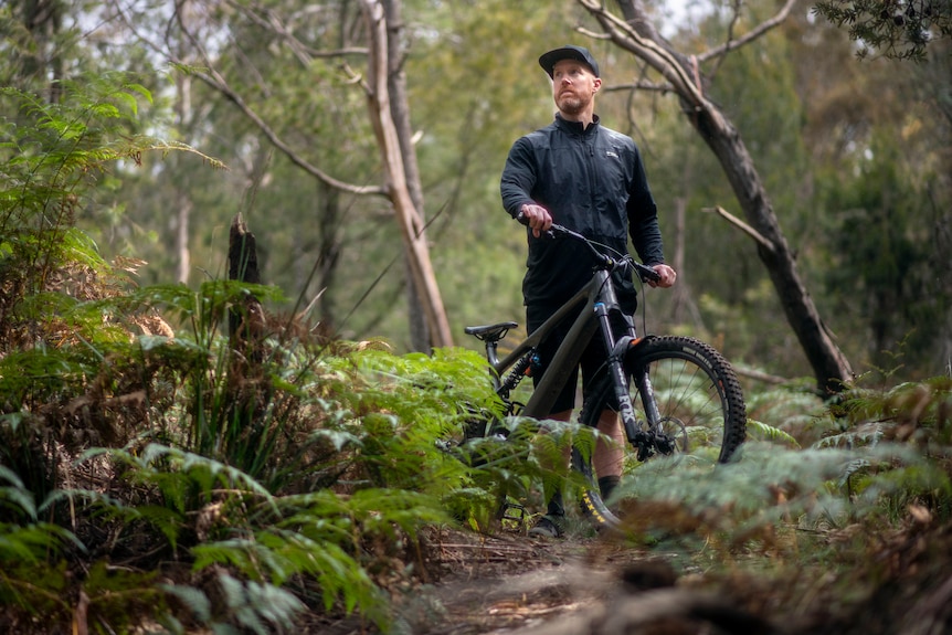 A man wearing a black cap steadies his mountain bike in a nature reserve surrounded by ferns.