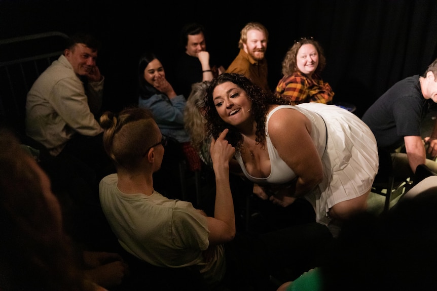 A woman in a white dress kneels in front of a man who points at her, with the audience grinning in the background