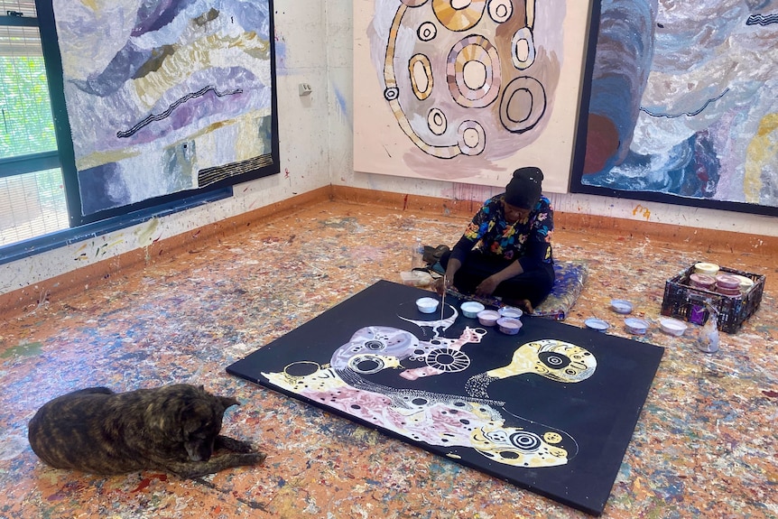 A dog sits peacefully beside her owner Pauline, who sits on the floor painting on a large canvas. 