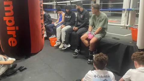 Simon Thake/BBC Young men sitting on the canvass of a boxing gym in sports clothes. Some have their hands strapped up.